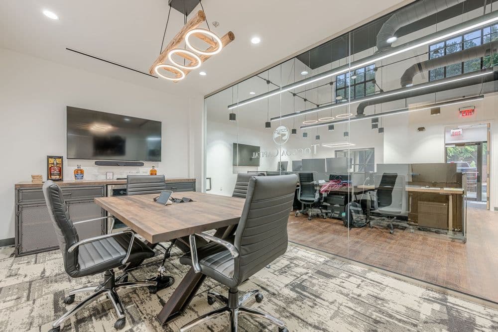 Modern conference room with a wooden table, leather chairs, and glass walls, featuring tech setup.