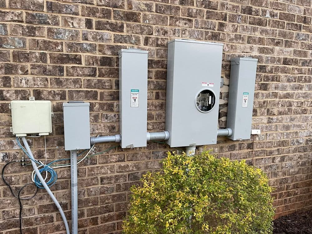 Electrical meters and service panels mounted on a brick wall with surrounding greenery.