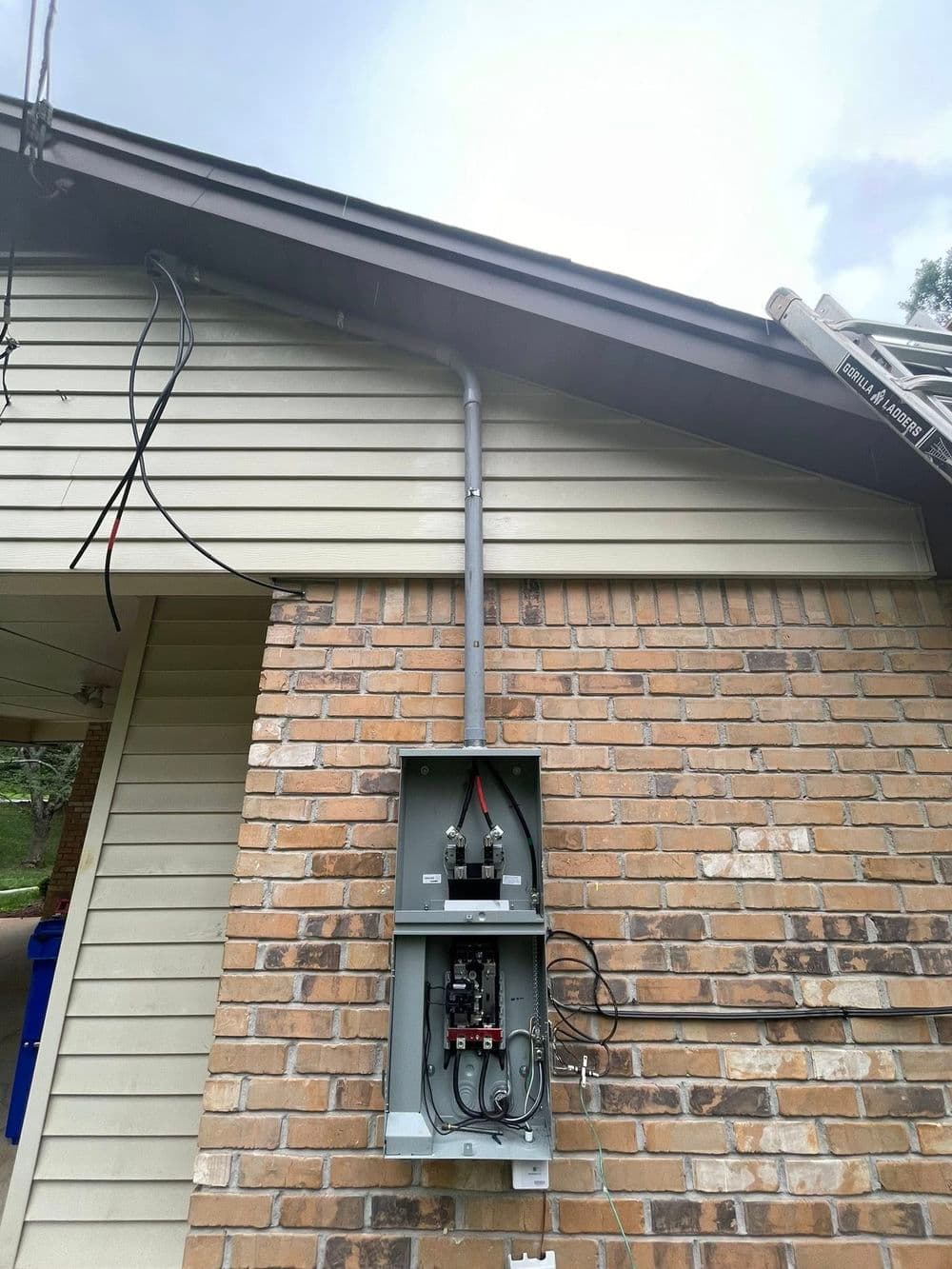 Residential electrical panel installation on brick wall, with utility wires and ladder nearby.